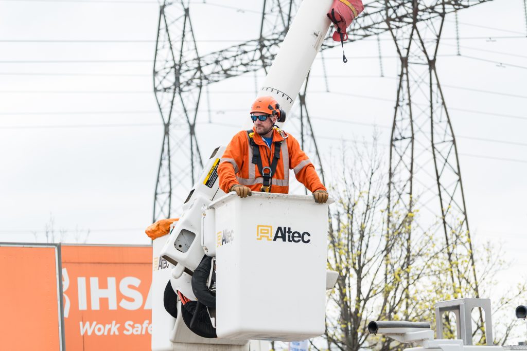 A man stands inside a aerial bucket wearing personal protective equipment with powerlines behind him.