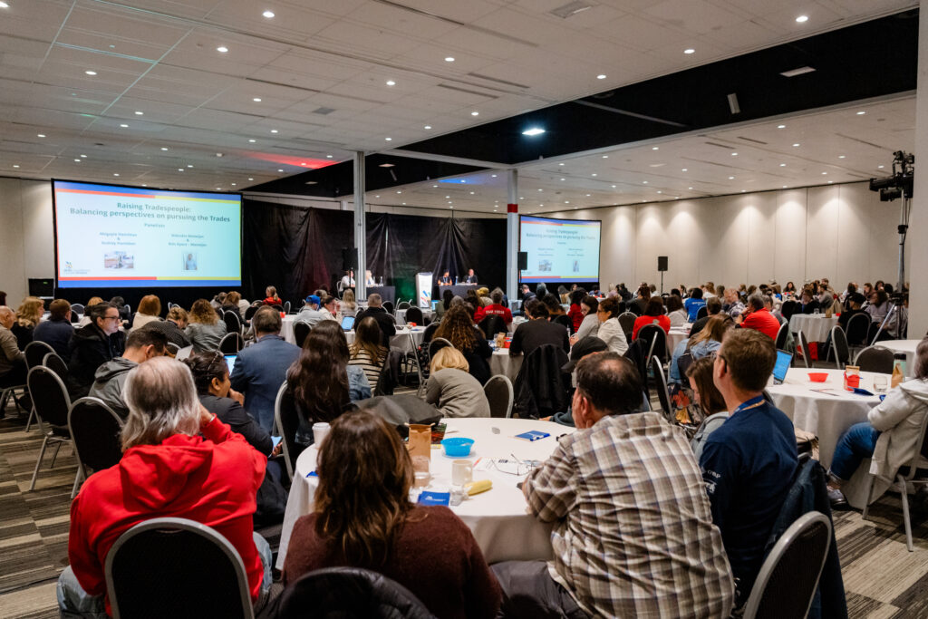 A photo of the backs of participants sitting at tables listening to four panel speakers on stage from the 2025 Skilled Trades Conference for Guidance Educators.